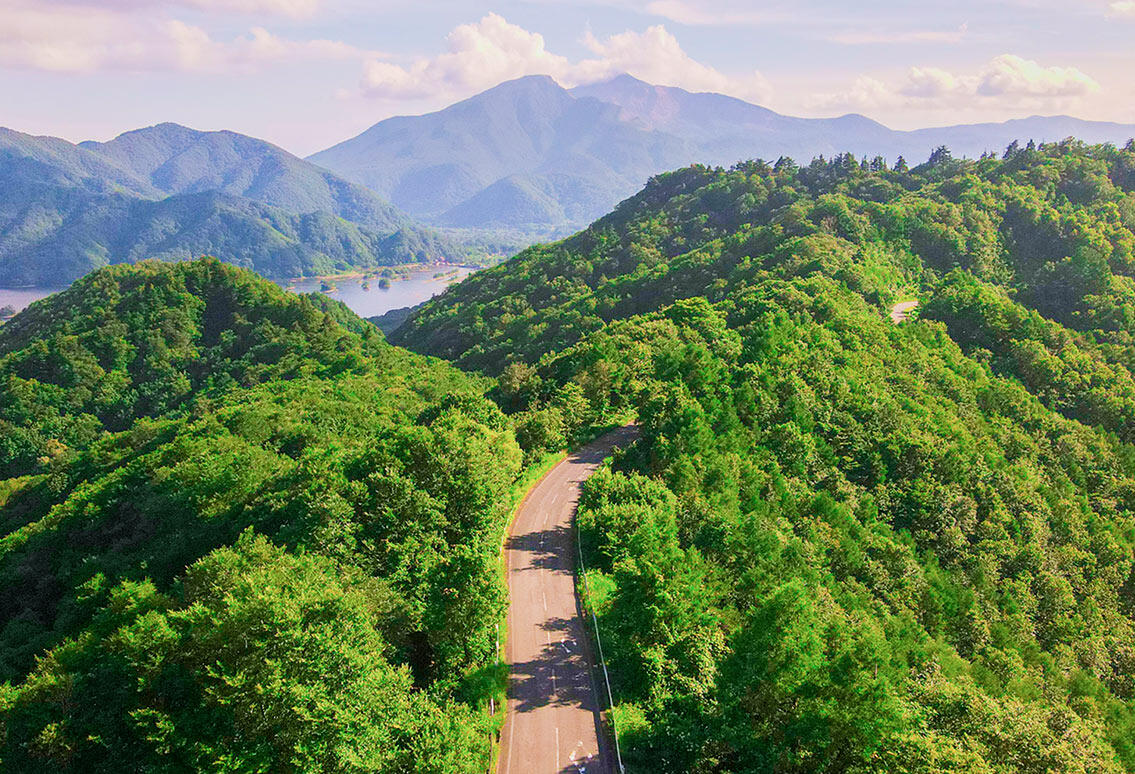 緑豊かな山々の尾根を縫うように道路が続く風景。遠方には檜原湖と磐梯山が望める。