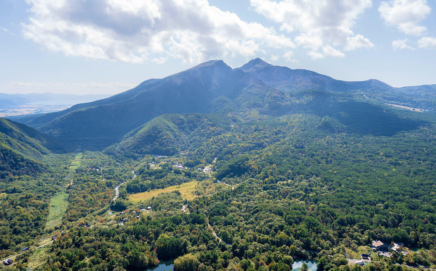 青空の下、深い緑の森と集落が広がる盆地を見下ろす風景。奥には双耳峰の磐梯山がそびえている。