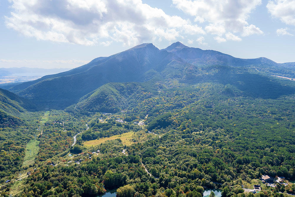 青空の下、深い緑の森と集落が広がる盆地を見下ろす風景。奥には双耳峰の磐梯山がそびえている。