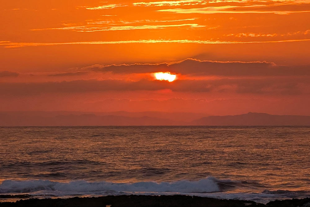 オレンジ色に染まった空と海の風景。雲の切れ間から太陽が沈み、手前には白波が打ち寄せている。