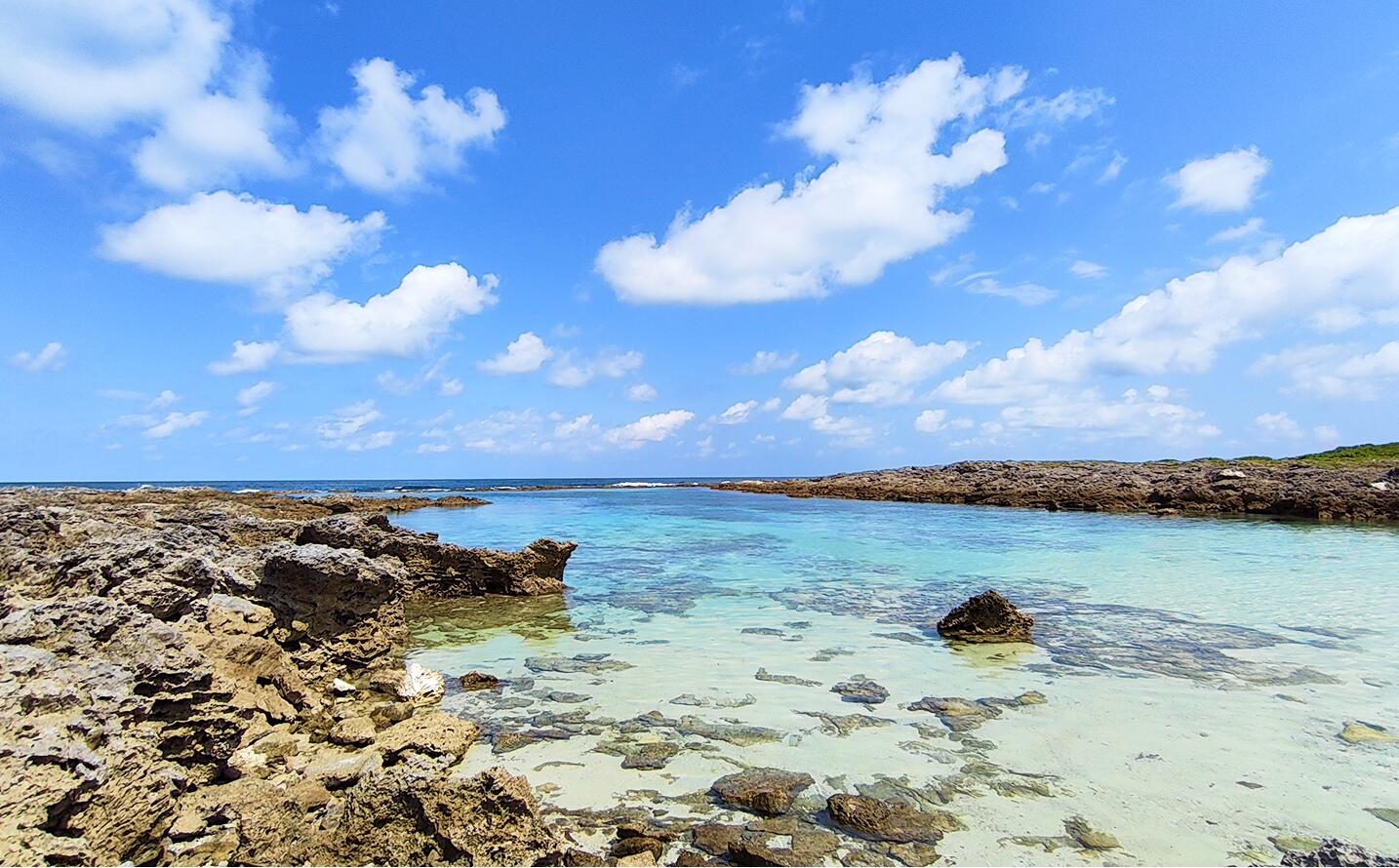 青空の下に広がる隆起サンゴ礁の海岸風景。透明度の高いエメラルドグリーンの浅瀬に岩礁が点在している。