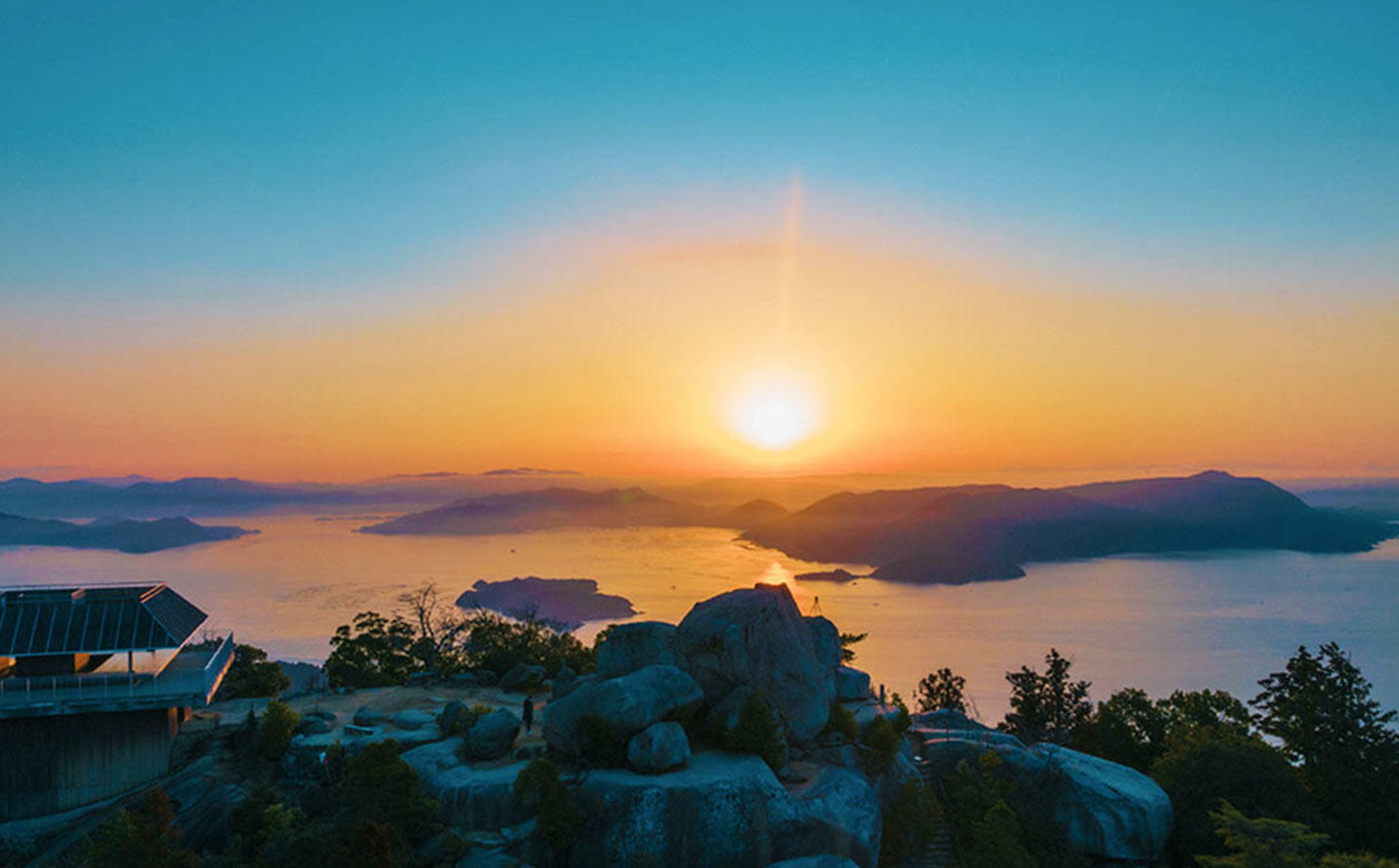 弥山山頂の展望台から望む瀬戸内海の夕景。手前に巨岩が連なり、水平線に太陽が沈んでいく。